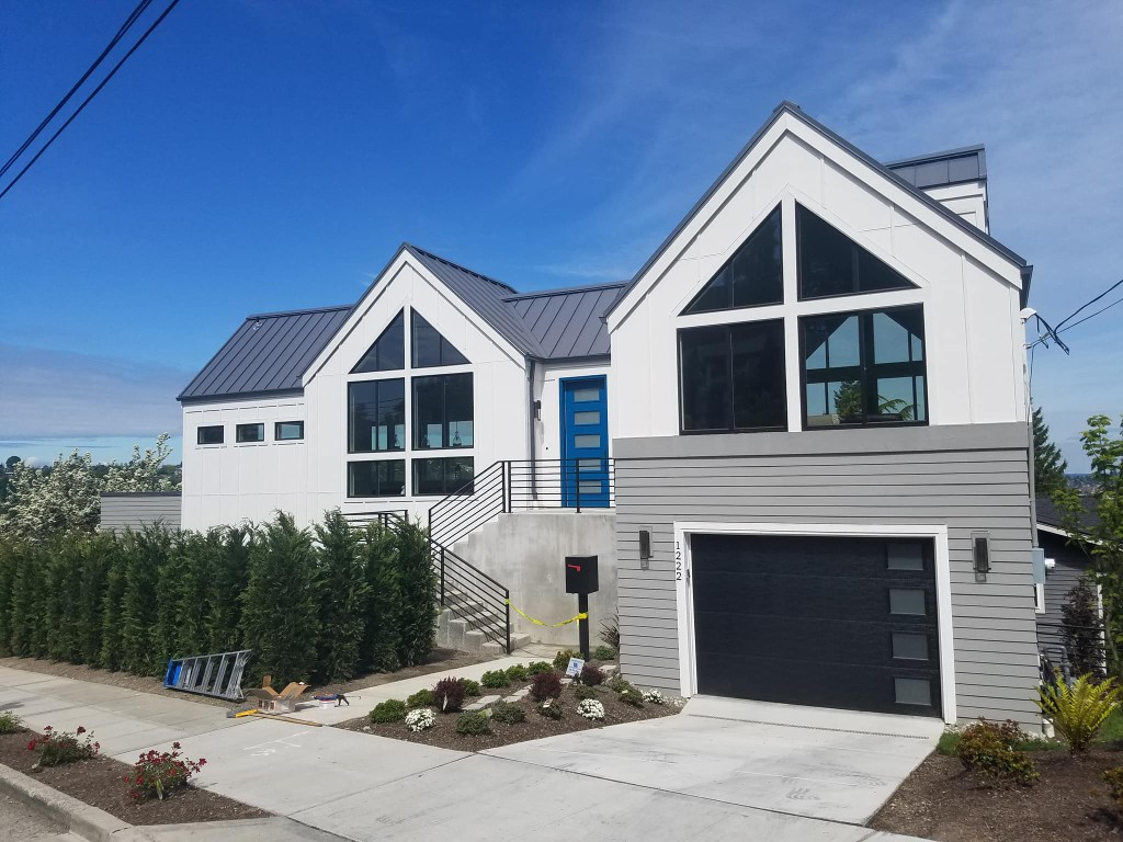 Modern two-story home with white siding, gray lower cladding, dark standing-seam metal roof, black window trim, and a bright blue front door.